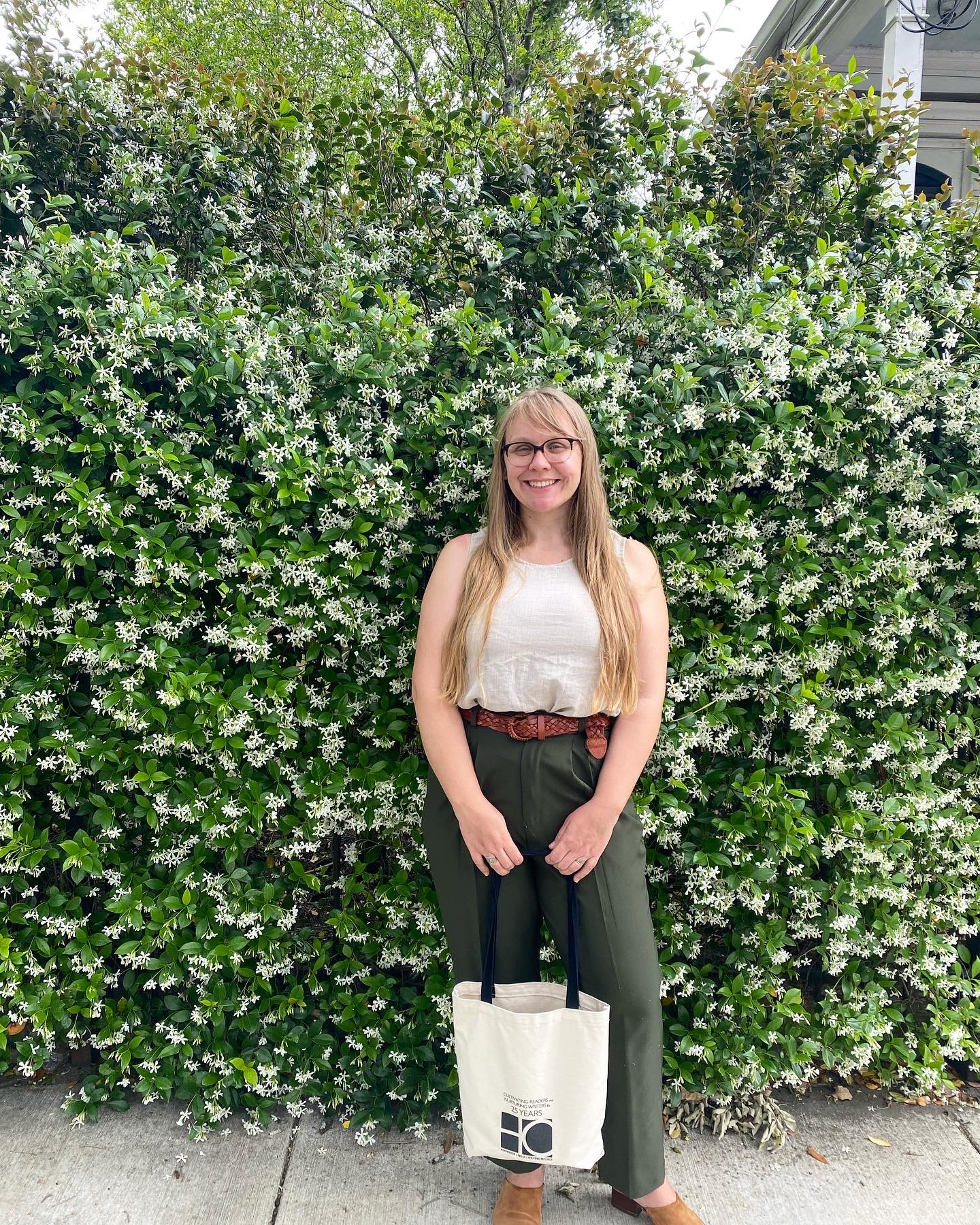 Image of poet Katherine Gaffney standing in front of a tall bush filled with flowers. Gaffney holds a canvas bag in both hands and smiles into the camera.