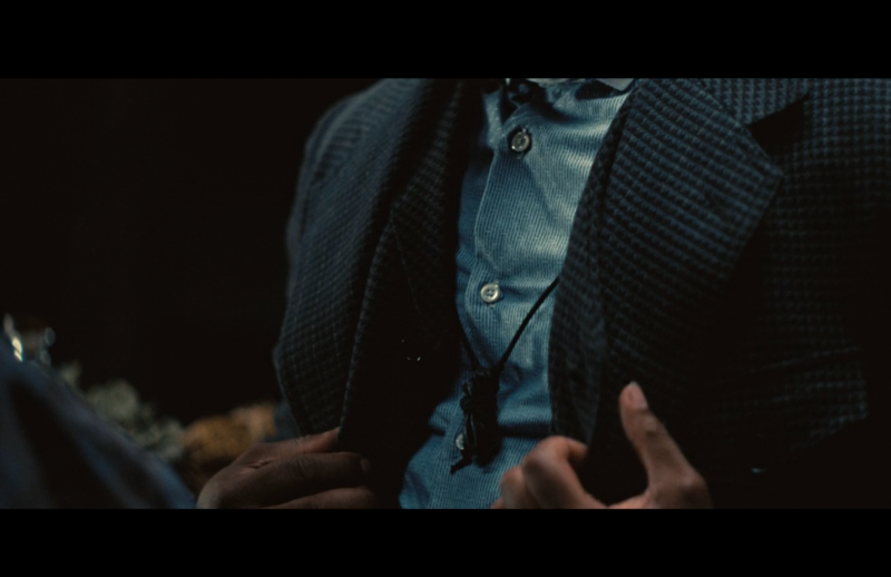 Smoke unbuttons his vest, revealing a black mojo bag secured by a string around his neck. Screen-capture by the author.